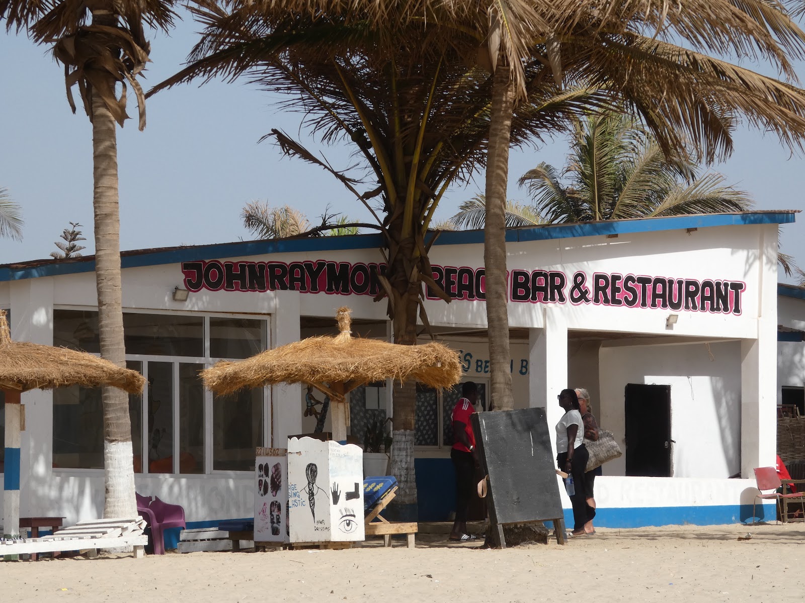 John Raymond's Beach Bar & Restaurant storefront with palm trees on Serrekunda beach
